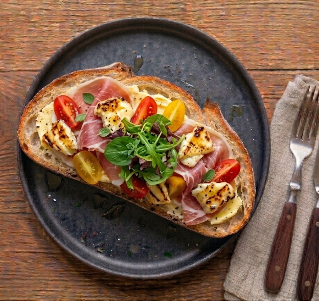 Tartine La Baiona signature — pain de campagne, beurre demi-sel, tranches de jambon de Bayonne, dés de tomme de brebis grillée aux bords caramélisés, tomates cerises rouges et jaunes, jeunes pousses de salade.