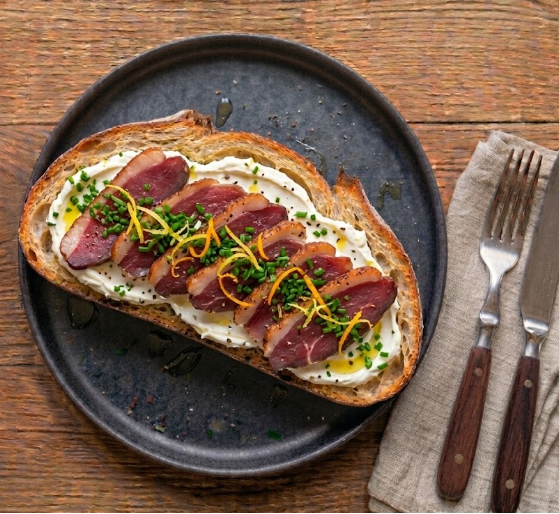 Tartine La Landaise signature — pain de campagne, couche de cream cheese, tranches de magret de canard séché à la chair rouge et liseré gras, ciboulette ciselée, zestes d'orange, filets d'huile d'olive.