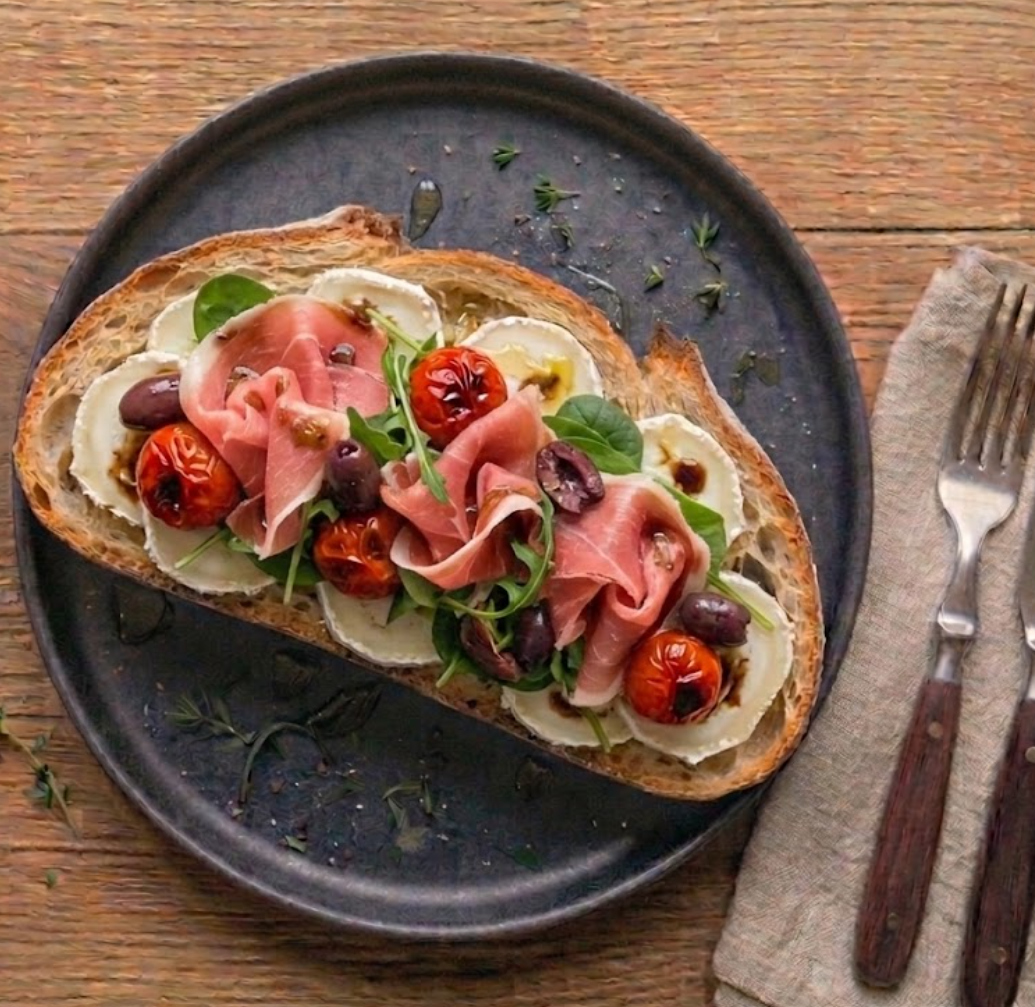 Tartine La Sully — pain de campagne, rondelles de chèvre frais, rosaces de jambon de Bayonne, tomates confites rouges, olives noires, jeunes pousses, filets d'huile d'olive.