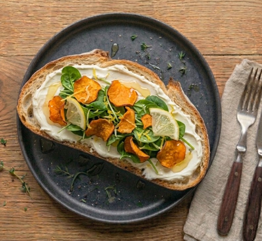 Tartine La Margaux — pain de campagne, couche généreuse de chèvre frais, pousses d'épinard, rondelles de citron, chips de patate douce orangées, filets de miel et huile d'olive.