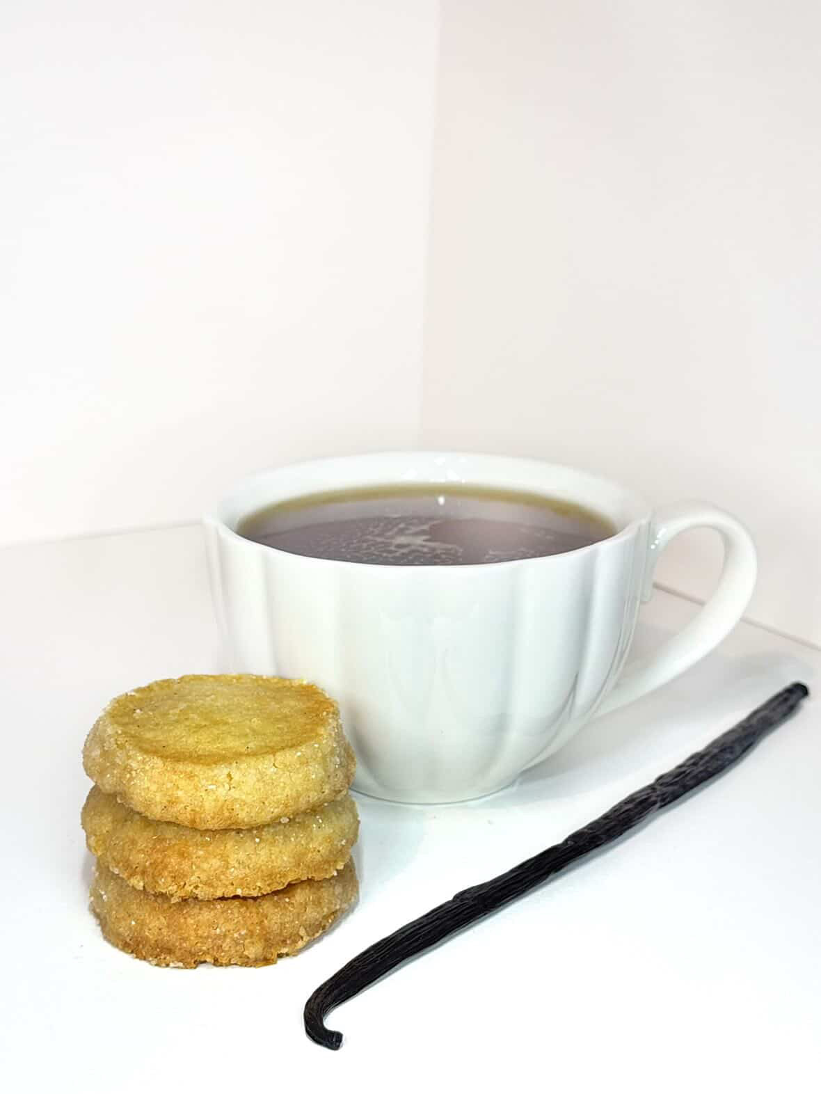 Thé chaud — tasse en porcelaine blanche, infusion ambrée, sablés vanille et gousse de vanille fendue à côté.